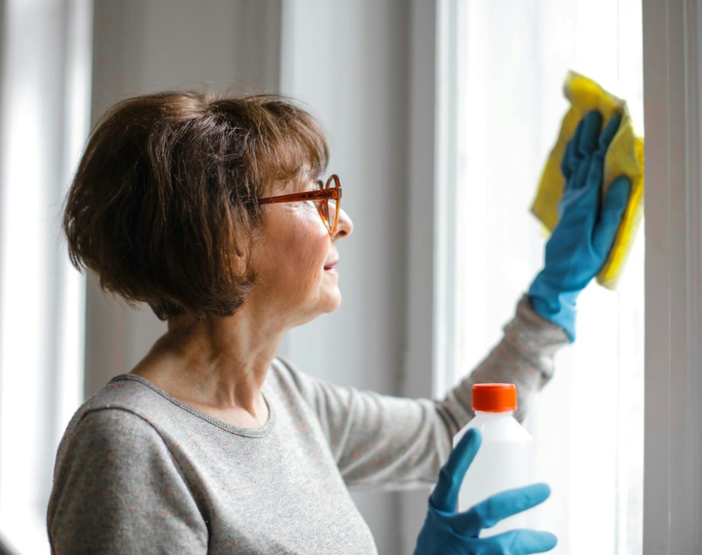 Elderly woman cleansing window indoors using gloves and cleaner.