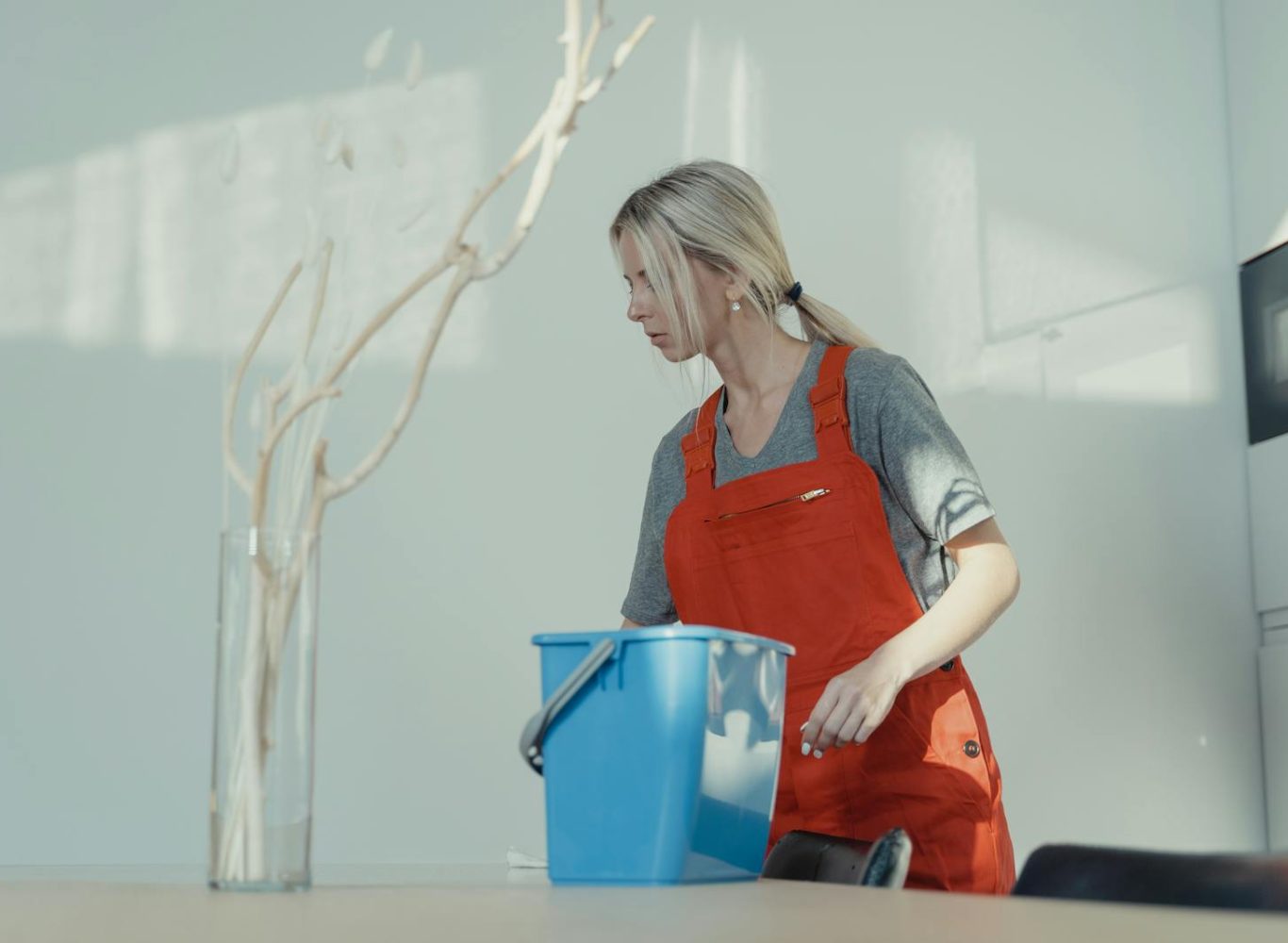 A woman in a red uniform cleans indoors with a blue bucket, ensuring a spotless environment.