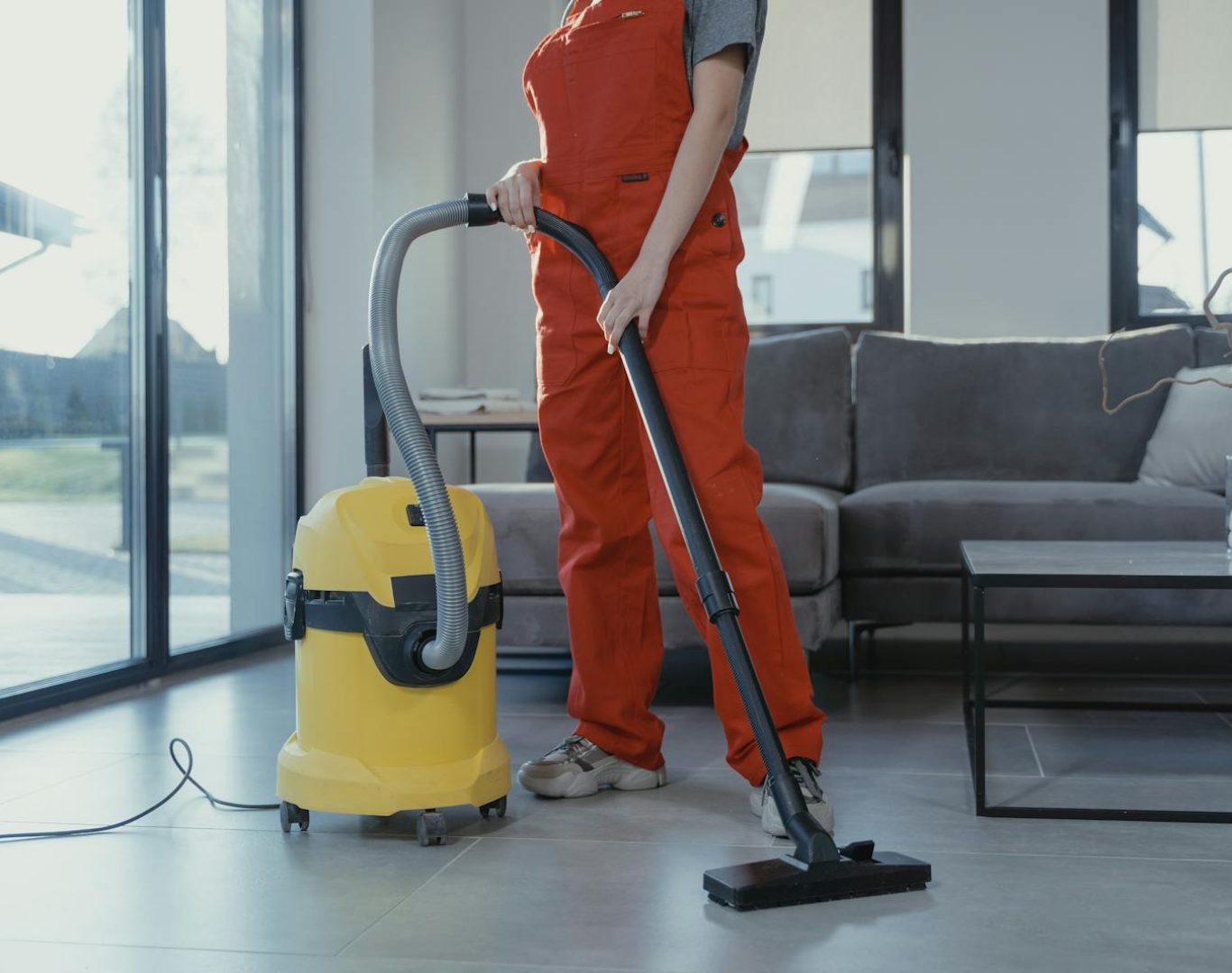 A professional cleaner in red coveralls vacuuming a modern living room with a yellow vacuum cleaner.